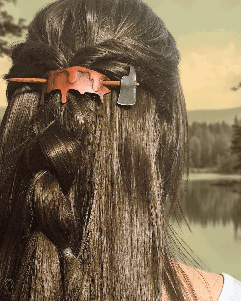 Vintage feeling photo of a person with a blood and axe shaped hair clip in their hair against a scenic background of a lake and trees.
