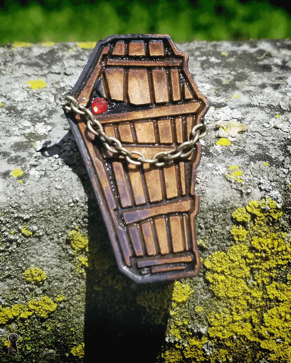 Wooden coffin-shaped lapel pin with a chain on a textured surface with greenery in the background on a bright day