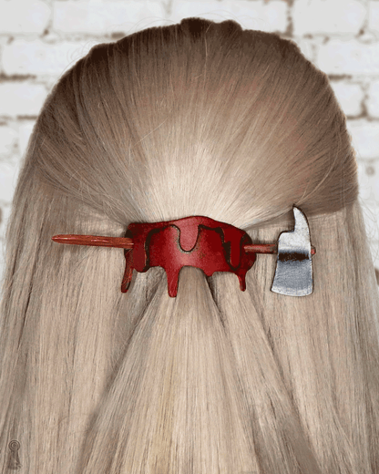 A blonde person with a blood and axe shaped hair clip in their hair against a white brick wall