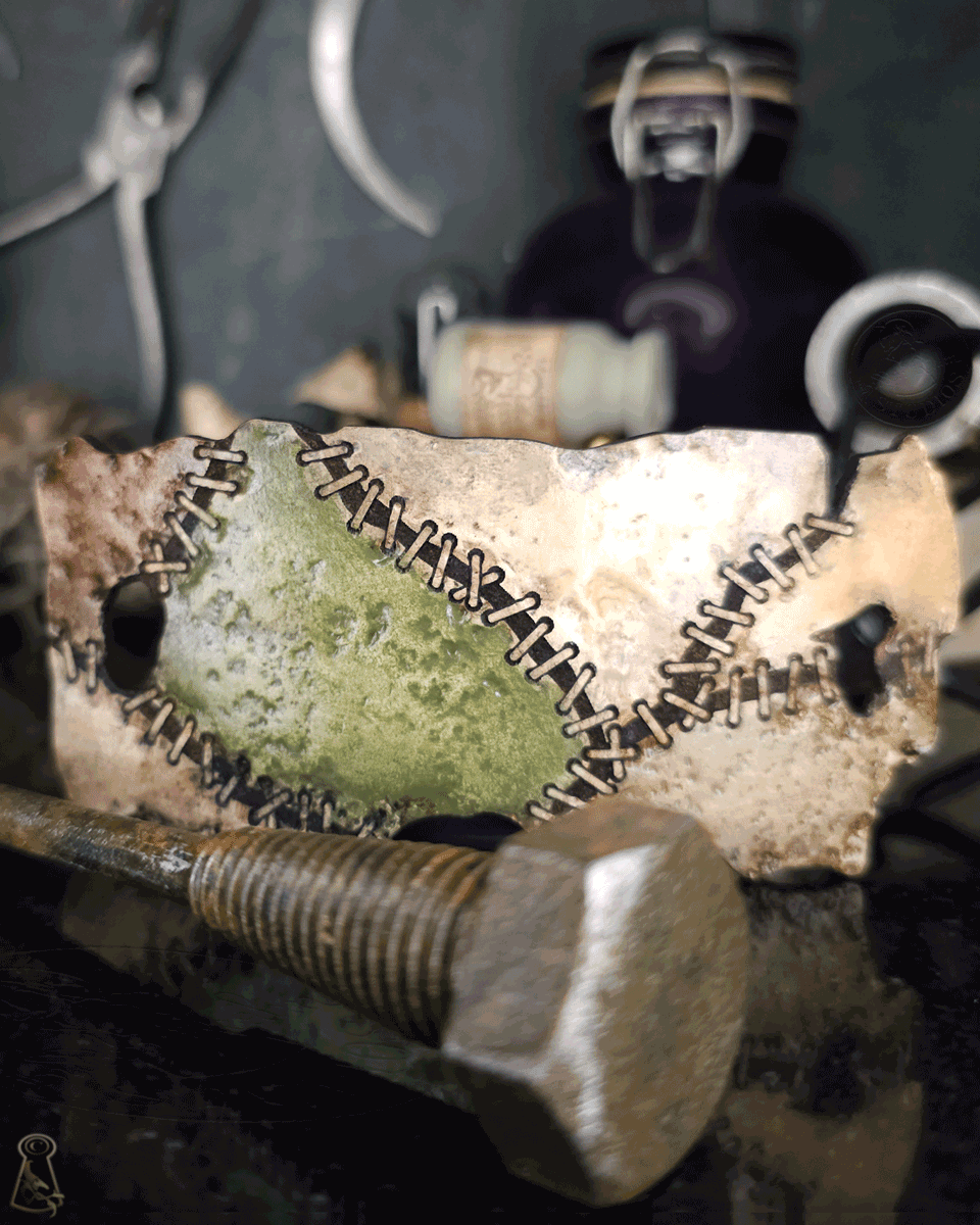 Close up of a metal finish bolt in front of a close up of a stitched monster skin patch on a dark background with jars and tools