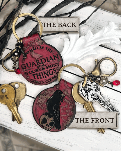 Front and back views of a red keychain design. A crow and skull on the front and text "So named by the goblin council: guardian of all spooky & shiny things" on a white background