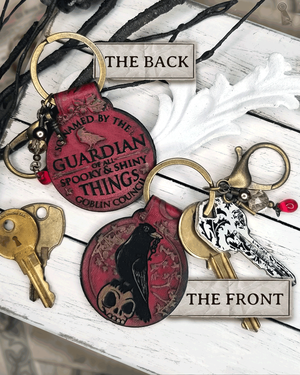 Front and back views of a red keychain design. A crow and skull on the front and text "So named by the goblin council: guardian of all spooky & shiny things" on a white background
