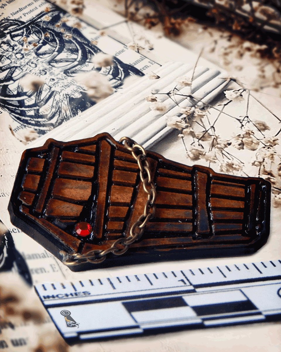 Wooden toe pincher coffin lapel pin with a red gemstone and a chain on a surface with measurements and decorative elements.