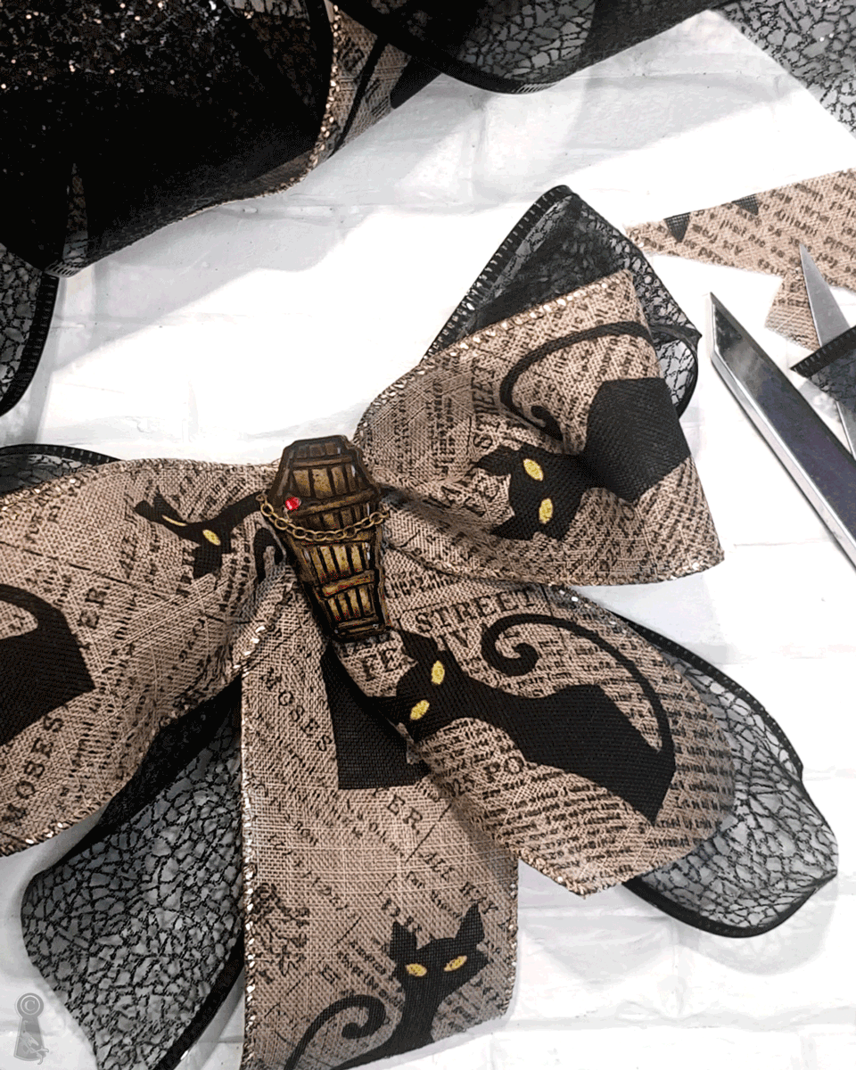 Decorative bow with black cats on a white background with a coffin charm pin in the center holding the bow together.
