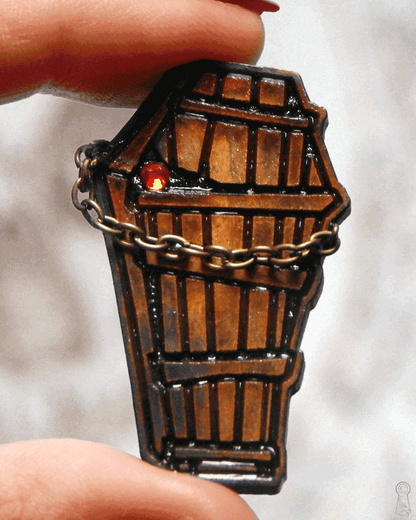 Close up of a wooden coffin-shaped charm pin with a red gen and bronze colored chain on a blurred background