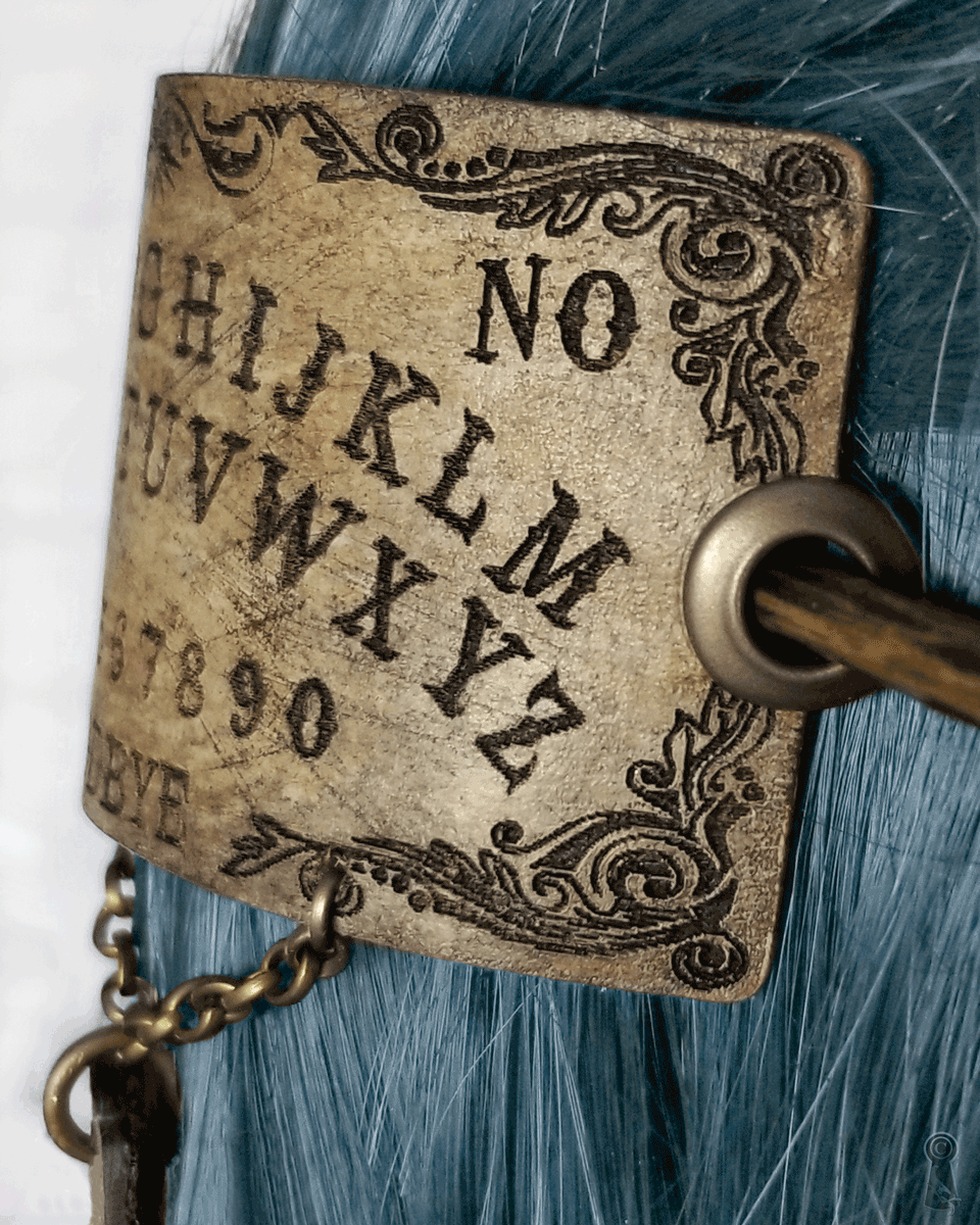 Decorative hairpin with Ouija board design against a teal blue hair.