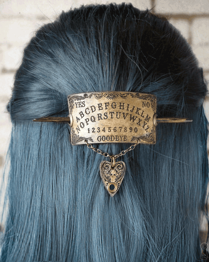 Hairpin with spirit board design and planchette charm on blue hair against a brick wall background
